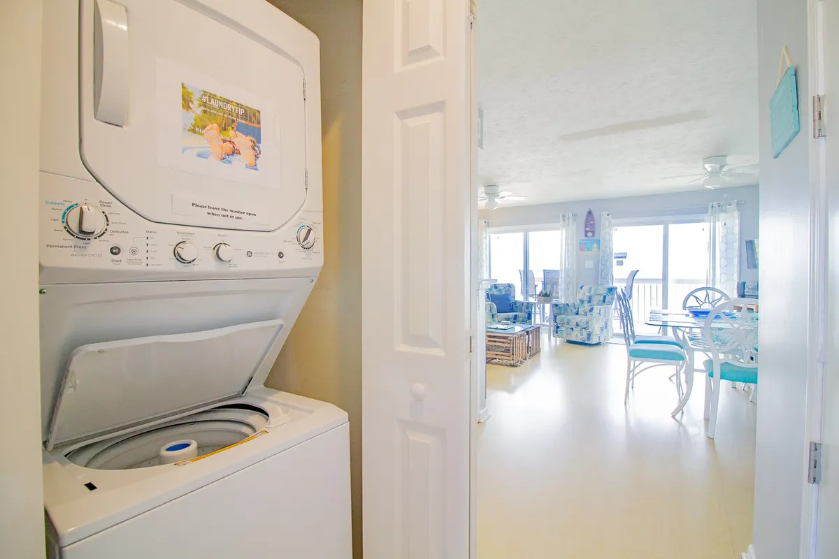 A washer and dryer are visible in a nook, with a portion of the living area and large windows in the background. The space is practical yet inviting, emphasizing the convenience of in-unit laundry for families or groups returning from beach adventures.