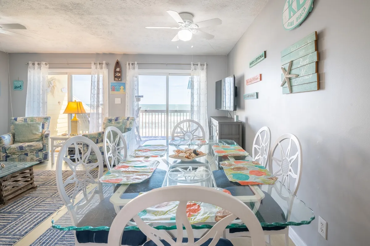 A spacious, open-plan dining and living area filled with natural light. The dining table is set with decorative plates and surrounded by comfortable seating, while the walls are adorned with beach-themed art. Large windows offer an inviting view of the beach, enhancing the airy, coastal atmosphere.