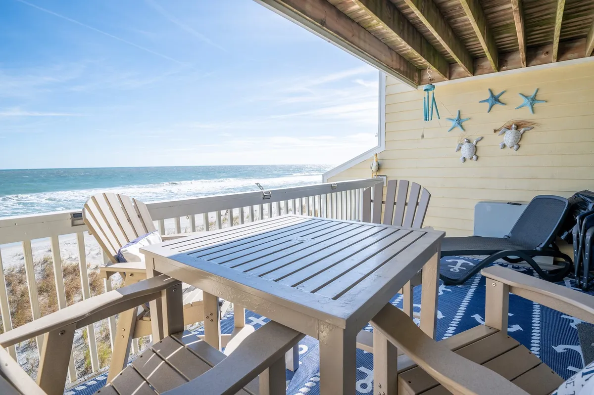 The image shows a serene oceanfront deck featuring a wooden table and classic Adirondack chairs positioned for guests to enjoy sweeping views of the Atlantic. Decorative starfish add a coastal touch, while the tranquil mood invites relaxation, dining, or socializing to the soothing sound of the waves.