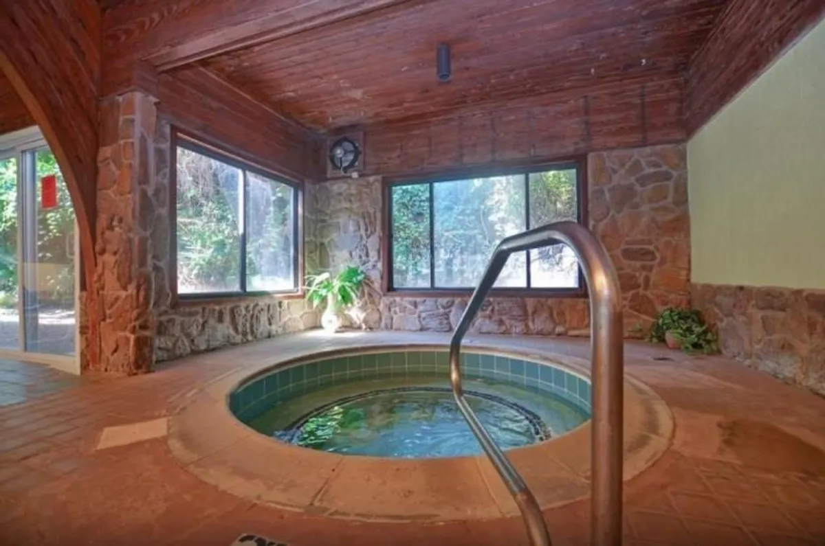 A tranquil indoor wellness area featuring a hot tub surrounded by natural stone walls, lush green plants, and large windows that let in natural light and hint at nature outside.