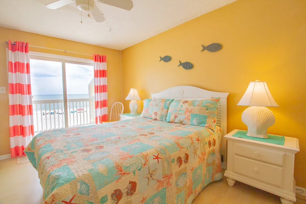 This image features a cozy bedroom with a bed covered in a colorful quilt, a window offering an ocean view, striped curtains, a shell-shaped lamp, and playful fish decorations. The atmosphere is tranquil and inviting, with soft lighting ideal for relaxing or reading.
