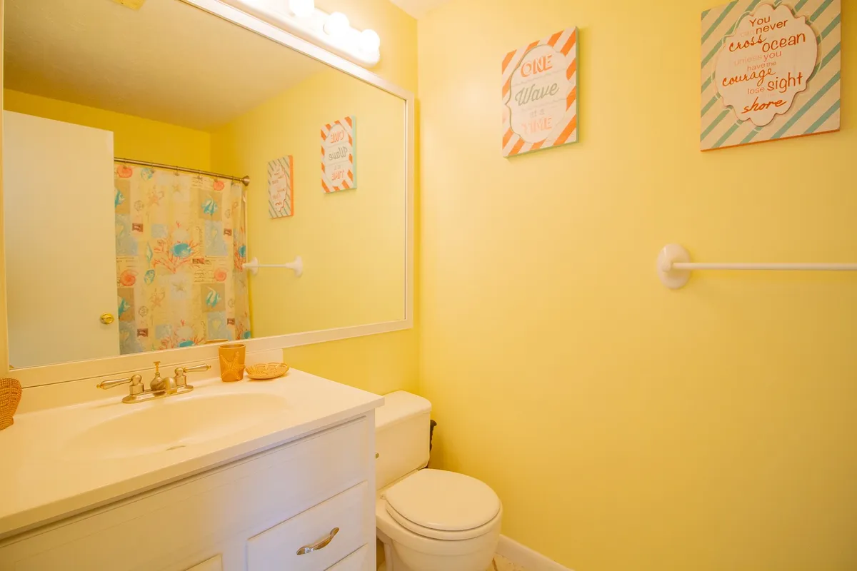 This bathroom features a large mirror, a colorful shower curtain, bright yellow walls, and decorative wall art. The room is playful and refreshing, offering a vibrant space for morning routines or relaxing after a beach day.