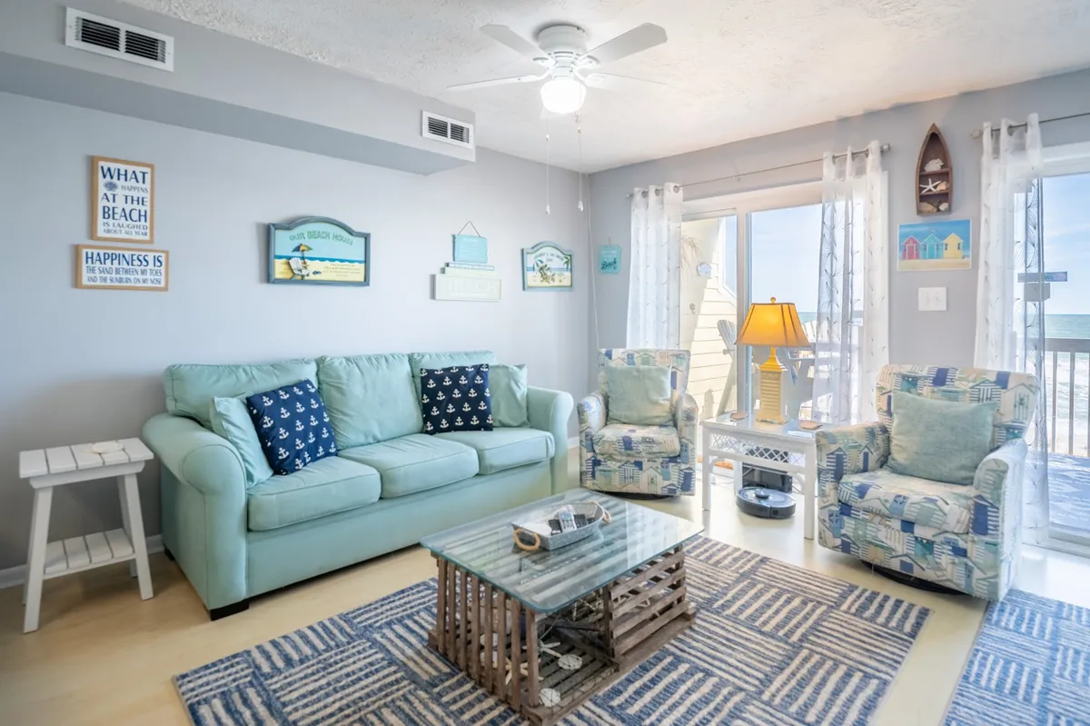 A light blue sofa, textured rug, glass-top coffee table, and beach-themed wall decor create a cheerful, tranquil living space. The scene invites relaxation and highlights private comforts inside the condo.