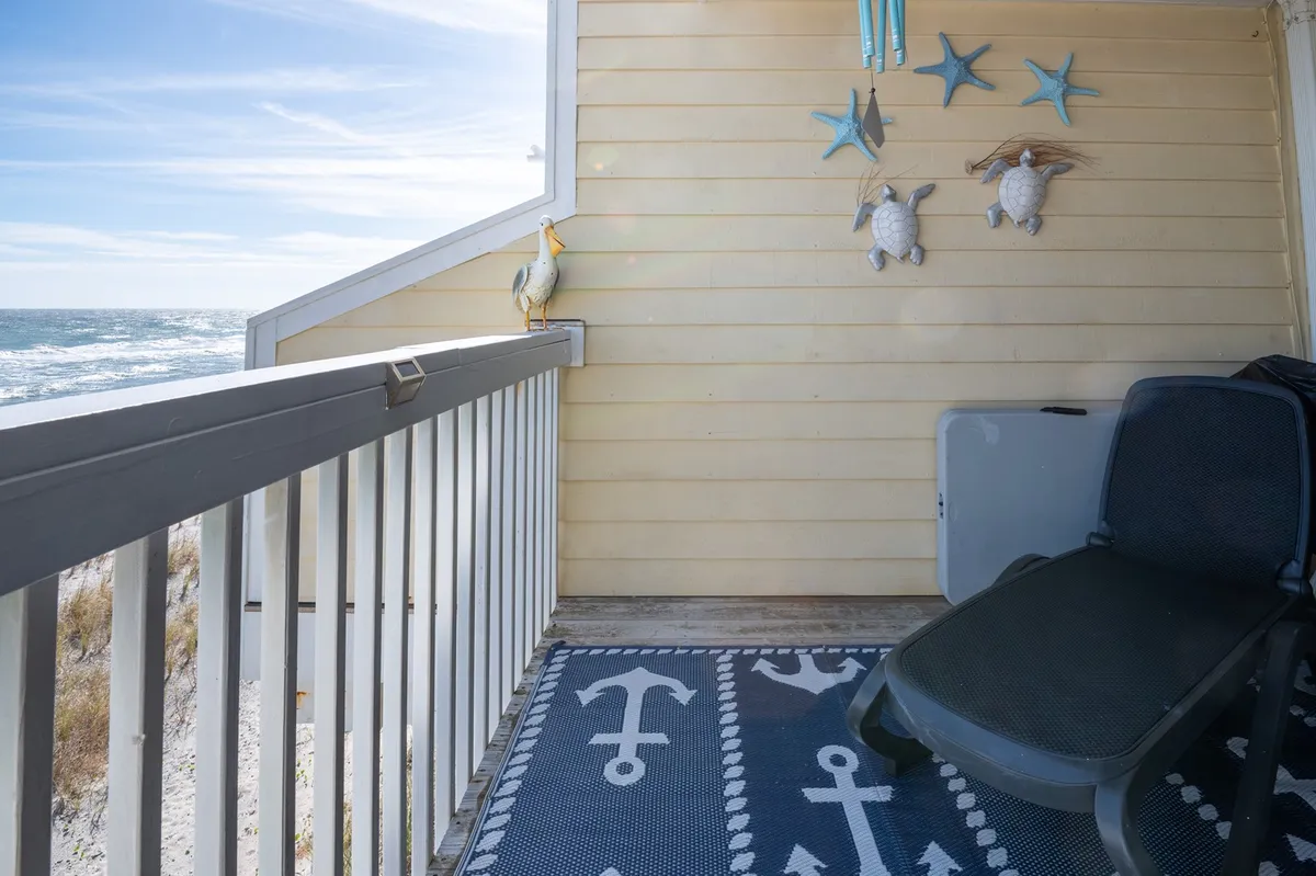 A sunlit private balcony with a lounge chair, decorative wall art, and a direct view of the beach and ocean. The area rug and cozy seating invite guests to relax while taking in the coastal scenery.
