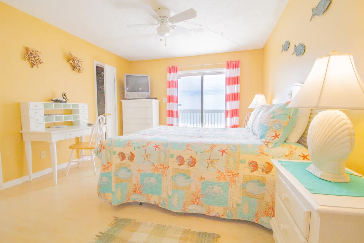 The scene shows a bright bedroom with a bed featuring a colorful quilt, a large window that frames an ocean view, a writing desk with a chair, and lamps on nightstands. The room is well-lit and inviting, emphasizing comfort and the surrounding coastal beauty.