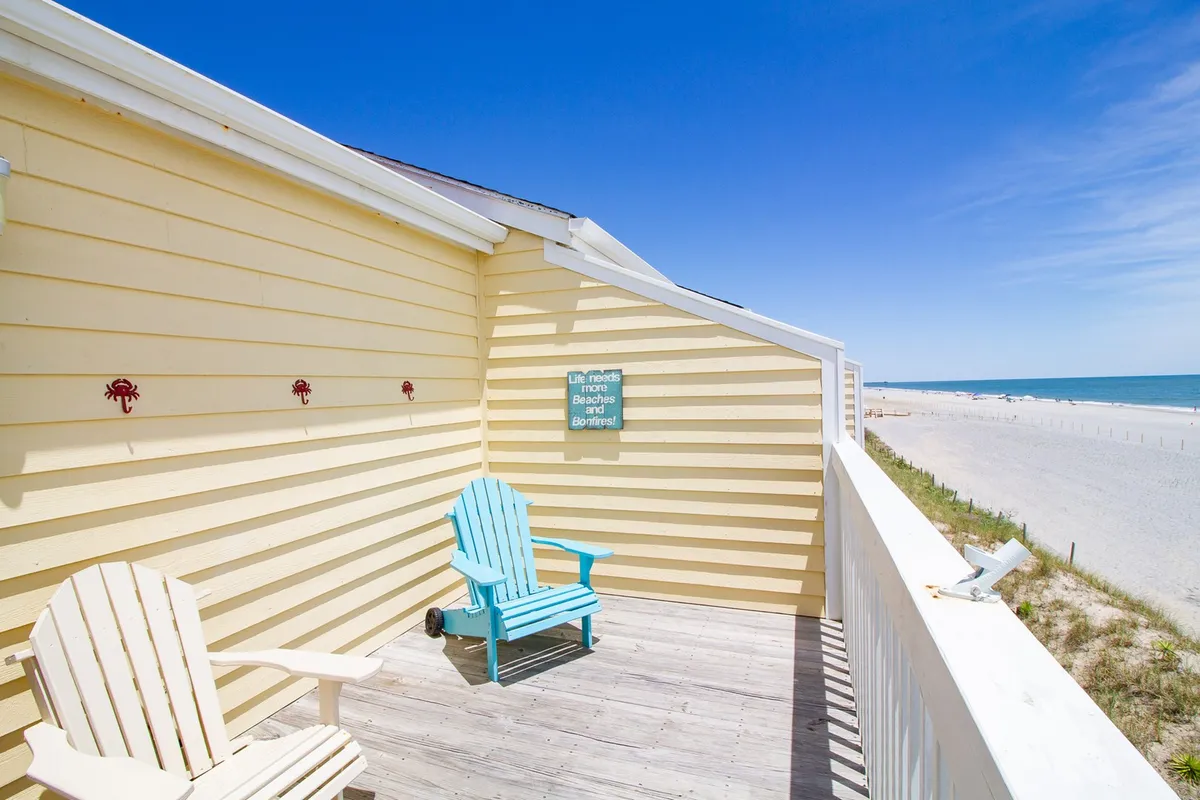 A tranquil deck with blue and white Adirondack chairs facing a beautiful beach view, complemented by decorative crab wall art and a motivational sign. The deck exudes relaxation, with space for socializing or enjoying drinks at sunset.