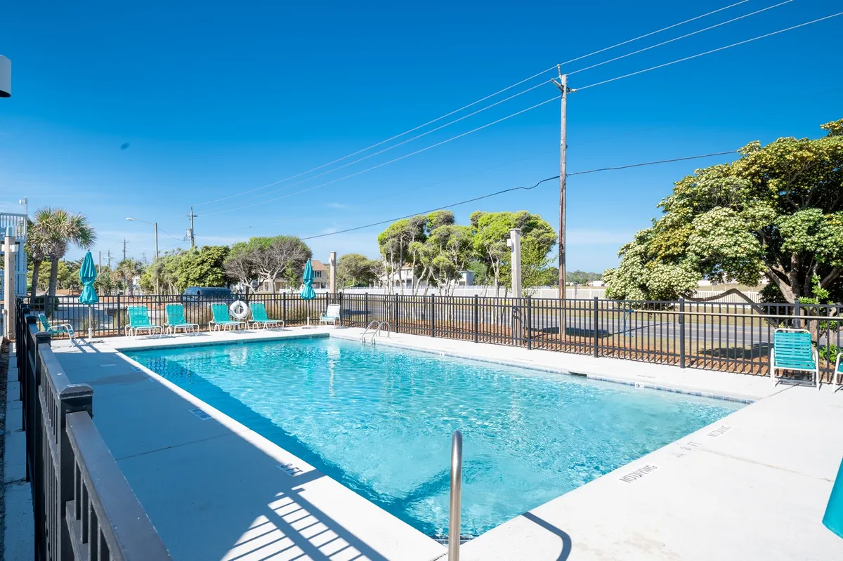 A sunlit outdoor swimming pool surrounded by poolside loungers, green trees, and a clear blue sky. The scene is lively yet relaxing, inviting guests to swim, sunbathe, or socialize in the communal area.