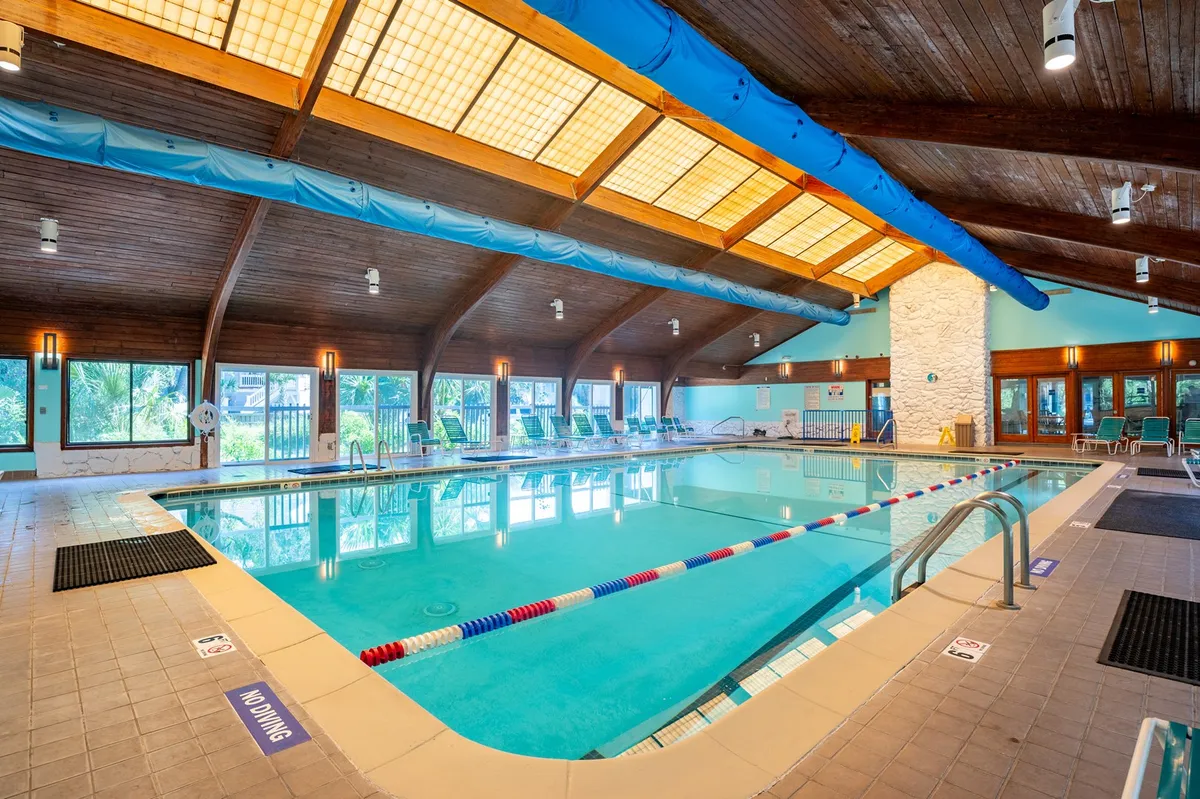 The indoor recreation center pool is depicted with tranquil blue water, wooden ceilings, and large windows that fill the space with natural light. Poolside lounge chairs invite guests to unwind, and the space exudes a calm, year-round retreat vibe.