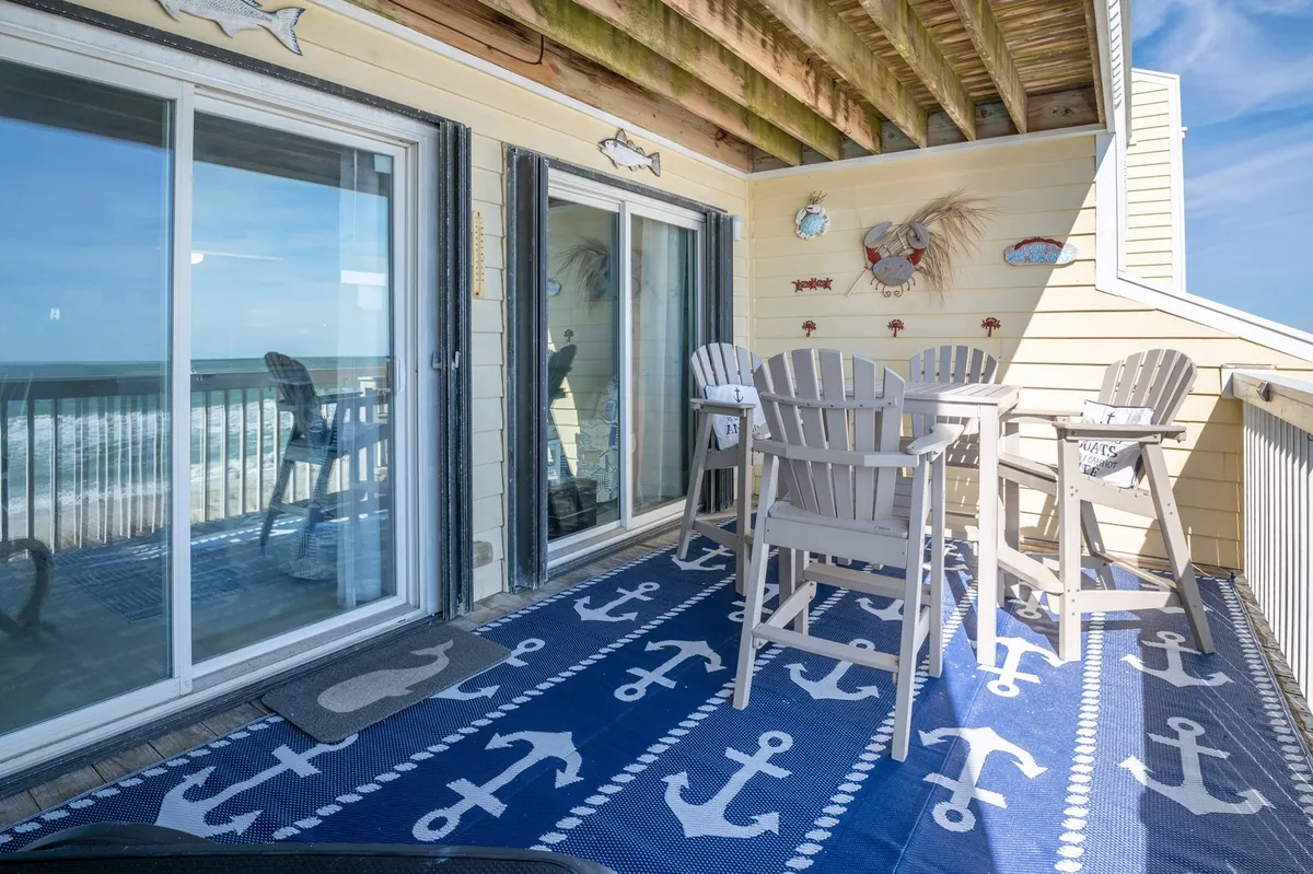 An inviting covered deck with comfortable outdoor seating overlooking the ocean. Decorative touches like wall art and an anchor-themed rug add a coastal flair, while the view promises tranquil mornings and stunning sunsets.